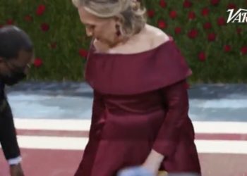 Die maskenlose Hillary Clinton läuft bei der Met Gala über den roten Teppich, während eine maskierte Dienerin sich um ihr individuelles Kleid kümmert (VIDEO)